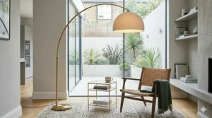 A statement arc floor lamp illuminating a minimalist sitting room with a woven armchair, set against large glass doors leading to a contemporary courtyard patio.